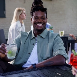 A man seated at a table in an outdoor covered area, with a cocktail in a lowball tumbler glass.