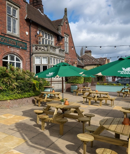 The exterior facade, signage, and beer garden seating area of the Golden Hind, with wooden picnic tables, shade umbrellas, and string lights.
