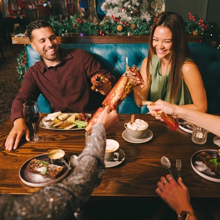 Four people sitting at a restaurant table, pulling Christmas crackers. A cheeseboard and two plates of Christmas pudding and custard sit on the table along with glasses of wine, a cup of coffee and a cup of hot chocolate, and Christmas decorations are visible in the background.