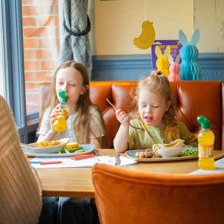 Two children sitting at an indoor table, wating a meal and drinking juice.