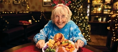 A roast dinner featuring roast meat, Yorkshire pudding, vegetables and gravy, served on a plate which sits on a table. A woman is sitting at the table, holding the plate with both hands and smiling at the camera.