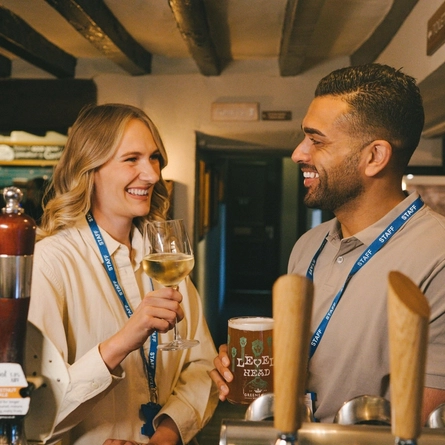 Two customers stood at the bar with a glass of white wine and a pint of Level Head at a Greene King venue.