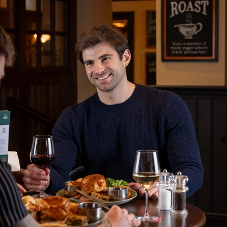 A person sits at a small round table in a pub-style restaurant, holding a glass of red wine. On the table are plates of roast dinner with Yorkshire puddings, vegetables, and gravy, along with condiments and an additional glass of white wine. Another person’s arm is visible in the foreground, reaching toward the food.
