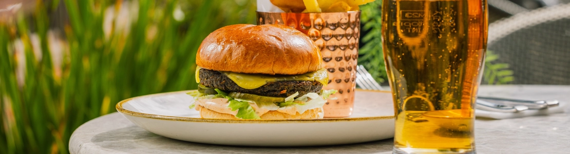 An advertising image of a plated Plant Based Cheeseburger and fries dish sat on a table within the exterior beer garden seating area at a Crafted venue.