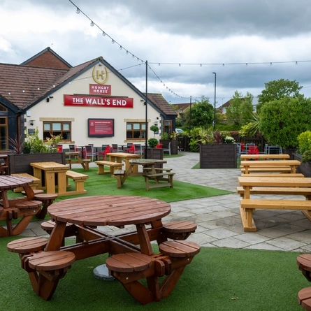 The exterior facade, signage, and beer garden seating area at The Wall's End in Wallsend, with wooden picnic tables, string lights overhead, and a TV on the wall.