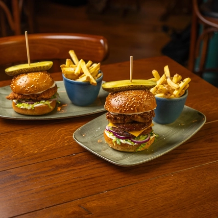 A Chicken Burger and a Pulled Pork Beef Burger, each served on a plate with a bowl of fries, sitting on a wooden restaurant table.