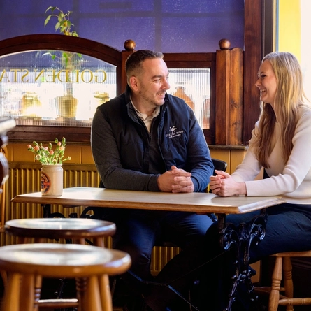 A Woman and Man sat at a table looking at each other smiling.