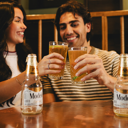 Two people sat in at a table in the interior seating area at a Crafted venue, holding two Modelo drinks into a 'cheers' position.