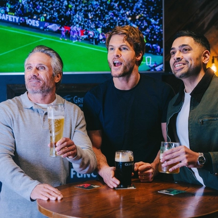 A group of friends stood round a table in the seating area in a Greene King pub with drinks watching sports.