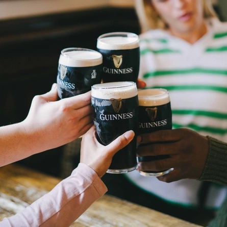 Four people are holding pints of Guinness beer, toasting at a wooden table. The focus is on the hands and the glasses.