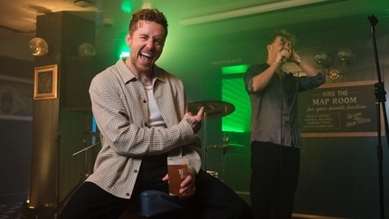 Two performers on a small pub stage during a live music event. One performer, Danny Jones, sits on a stool holding a pint glass with visible “Hazy Day” branding and gestures towards the singer standing behind with a microphone. Green stage lighting, a drum kit and pub signage create a lively live entertainment setting.