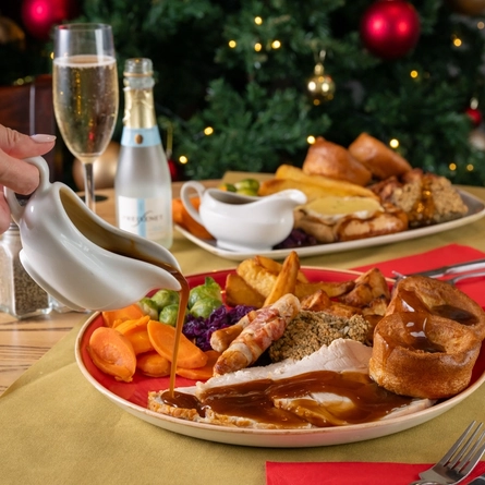 A hand holding a gravy boat, pouring gravy over a plated turkey roast dinner.  The table is decorated in a festive manner.