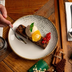 An overhead view of a plated portion of Christmas Pudding topped with ice cream. on a festive decorated wooden table.  A hand holding a spoon is about to take a piece. 
