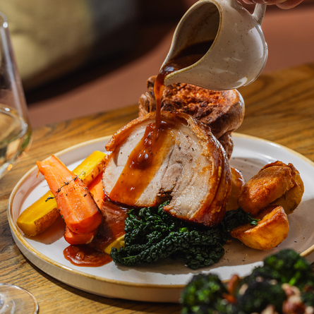 A lifestyle image showing a jug of gravy being poured over a plated Roast Pork Loin dish sat on a table within the interior restaurant seating area at The Four Oaks.