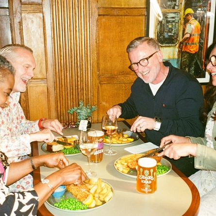 A group of friends sat in the interior restaurant and seating area in a Greene King London venue, enjoying served mains of fish and chips with drinks.