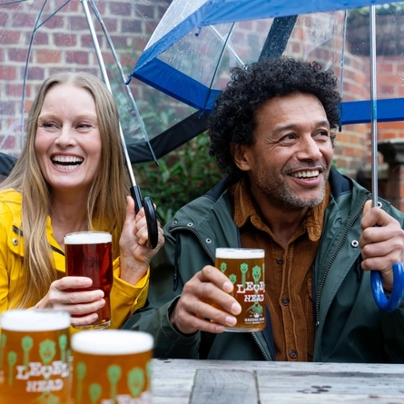 Two people enjoying beers in the rain under an umbrella.