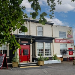 The exterior facade, signage, and seating area of The Cherry Tree in Newcastle-Under-Lyme; with shade umbrellas on the tables, and string lights hanging above the seating area.