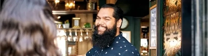 A man in a patterned blue shirt smiling at someone in the doorway of a pub.