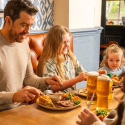 People sit at a wooden table in a pub dining area, eating a roast meal with meat, vegetables, potatoes, and Yorkshire puddings. Pints of lager and bottled juice drinks are on the table