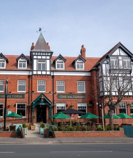 A view from across the road of the exterior facade, signage, and beer garden seating area of The Victoria in Lytham St Annes, with shade umbrellas on some of the tables.