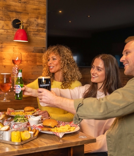 An image showing a group of people sat around a table enjoying mains and sharer dishes with various drinks, celebrating with a cheers at a Flaming Grill venue.