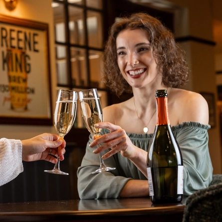 Two women seated at an indoor table.  They are both holding champagne flutes containing prosecco in a toast.  A bottle is on the table between them.