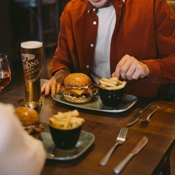 Person seated at a pub table reaching for chips, with a bacon cheeseburger and pint of Guinness in front.