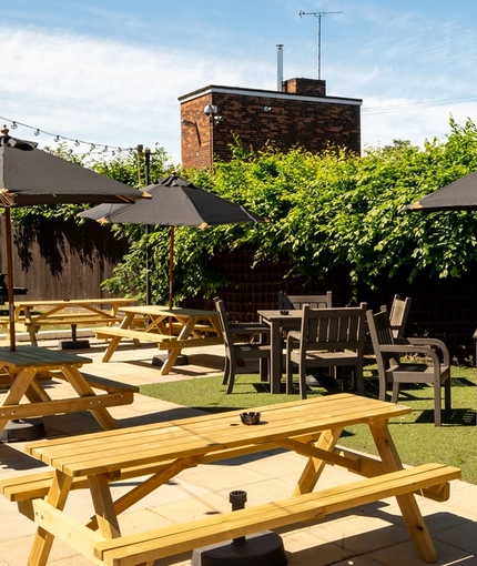 The exterior beer garden seating area at The Bramford Arms in Woodsetton, with paved and grass areas, wooden picnic tables, shade umbrellas, and string lights.