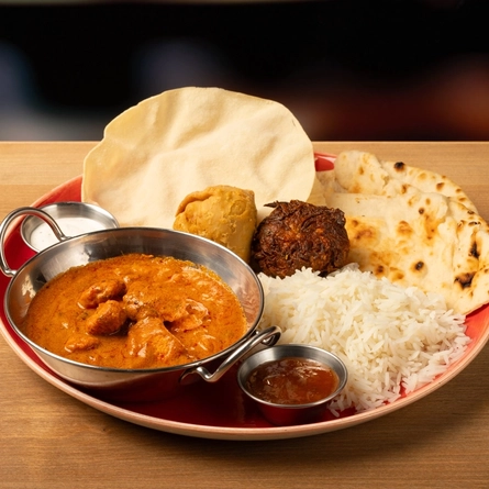 A Balti dish of curry on a plate with rice, naan bread, poppadom and a dipping sauce  on a wooden table.
