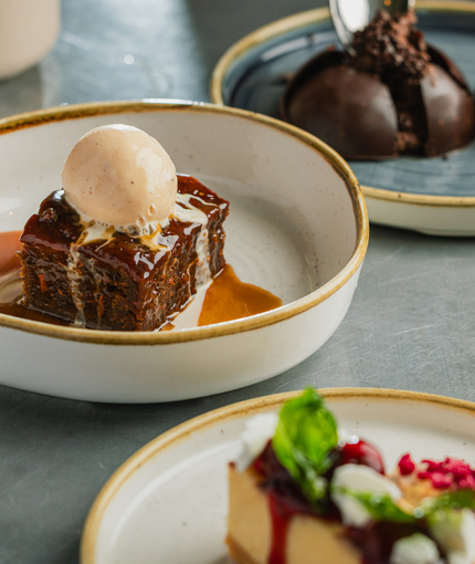 A lifestyle image showing a person sat at a table enjoying a Dark Chocolate Mousse Bomb Dessert with 2 Dessert dishes also seen in the image including Sticky Toffee Carrot Cake Pudding & Strawberry Shortbread Cheesecake at a Crafted venue.