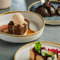 A lifestyle image showing a person sat at a table enjoying a Dark Chocolate Mousse Bomb Dessert with 2 Dessert dishes also seen in the image including Sticky Toffee Carrot Cake Pudding & Strawberry Shortbread Cheesecake at a Crafted venue.