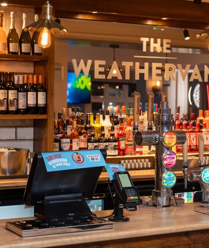 A close up view of the bar at The Weathervane, with bottles of wine and alcoholic spirits on wooden shelves behind the counter.