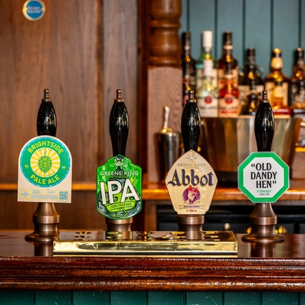 A close up view of two golden drinks trays and the beer taps for Brightside Pale Ale, Greene King IPA, Abbot Ale, and Old Dandy Hen; on the bar at the Phoenix.