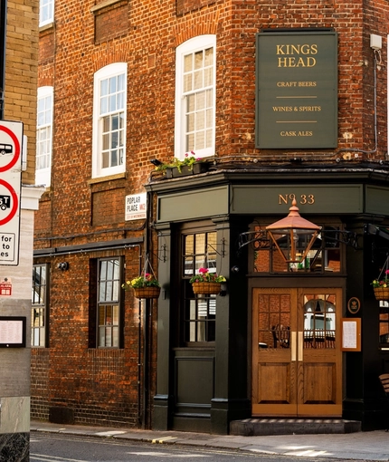 The exterior facade, signage, and seating area at the King's Head in Bayswater, with double wooden doors; a dark green awning hanging over the wooden tables and chairs; flower baskets hanging on the walls; and flower boxes beneath the upper windows.