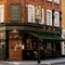 The exterior facade, signage, and seating area at the King's Head in Bayswater, with double wooden doors; a dark green awning hanging over the wooden tables and chairs; flower baskets hanging on the walls; and flower boxes beneath the upper windows.