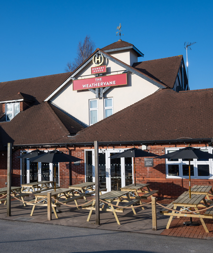 The exterior facade, signage, and seating area at The Weathervane in Stoke On Trent, with wooden picnic tables, shade umbrellas, and string lights.