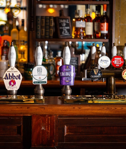 The interior bar focusing on a pump with several pump clips including Peroni, Estrella, Level Head, Hazy Day, Green King IPA, Abbot, Three Jolly Butchers Pale Ale, Tribute, Guinness, Aspall Cyder, Camden Hells and Birra Moretti all served at the Three Jolly Butchers.