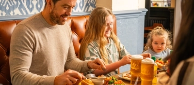 People sit at a wooden table in a pub dining area, eating a roast meal with meat, vegetables, potatoes, and Yorkshire puddings. Pints of lager and bottled juice drinks are on the table
