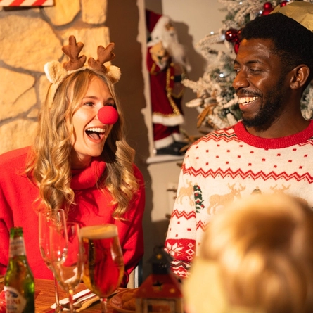 Two people in festive jumpers sit at a Christmas dining table in a pub decorated with a snow-covered Christmas tree and seasonal decorations. One person wears a reindeer antler headband and a red novelty nose, while the other wears a paper crown from a Christmas cracker. Glassware and drinks are arranged on the table, creating a fun and festive Christmas Day gathering.
