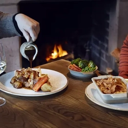 A man and a woman with plates of food in front of them, pouring gravy onto their meals.