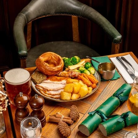 A plated turkey dinner on a festive decorated wooden table with a tankard of beer, a glass and bottle of sparkling wine and a stemmed glass containing a spirit cocktail.