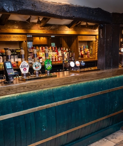 A close up view of the bar at The Fox & Hounds in Bursledon, with wooden beams, bar stool seating, and bottles of wine and alcoholic spirits on wooden shelves behind the counter.