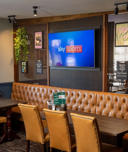 An interior restaurant seating area at the Rudds Arms; with a long, upholstered booth seat; and a large TV, framed artwork, and artificial plants on the wall above it.