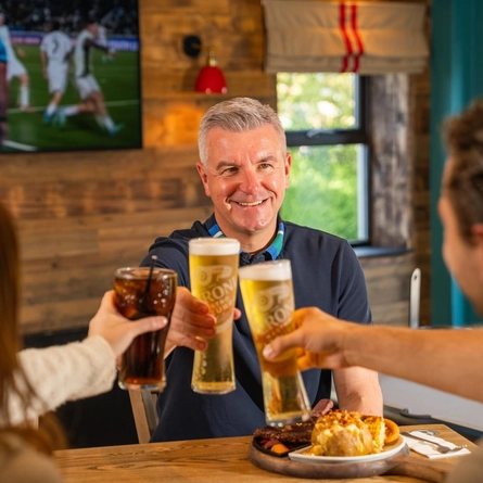 A image focused on a Dad holding a pint into a cheers position, smiling with children seated opposite on Fathers day at a Flaming Grill venue.