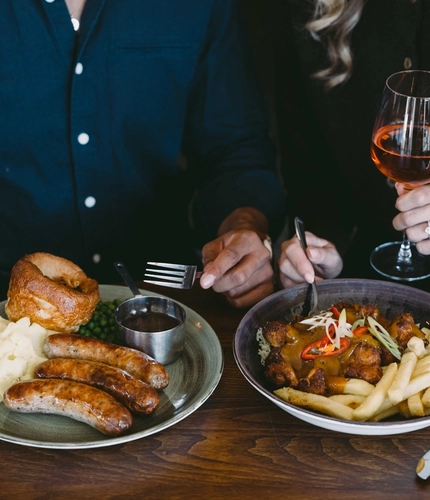 Two people seated together at a pub table with plates of sausages, mash and Yorkshire pudding, and curry with rice and chips, alongside a pint of beer and a glass of rosé.