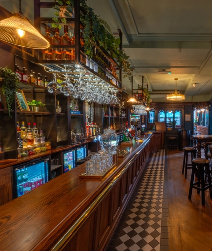 The wood panelled bar and interior restaurant seating area at The Plough, with glasses hanging in racks above the counter, bar stool seating, and framed artwork on the wall.