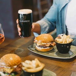 Bacon cheeseburger in a sesame-seed bun with red onion and mayonnaise, served with chips as a person holds a pint of Guinness.