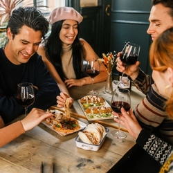 friends sitting around a table with red wine and a variety of side dishes spread across the table  