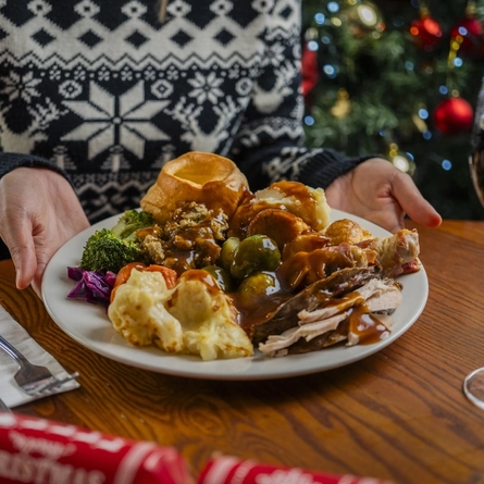 A close up view of a plate of carvery roast dinner sitting on a wooden table next to cutlery, a Christmas cracker, and a glass of red wine. A person sitting at the table is holding the plate and a Christmas tree is visible in the background.