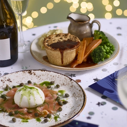 An advertising image showing 2 plate New Years Eve dishes and a glass of sparkling white wine sat on a table within the interior restaurant seating area at a Chef & Brewer venue.